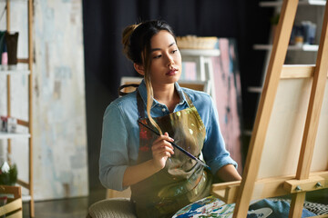 An Asian artist in an apron stands in her studio, thoughtfully considering her next brushstroke on a canvas.
