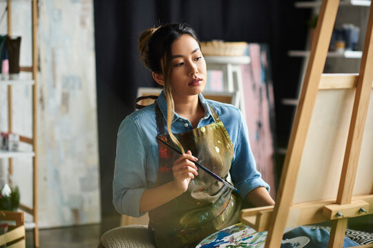 An Asian artist in an apron stands in her studio, thoughtfully considering her next brushstroke on a canvas.