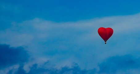 Fototapeta premium A single, red, heart-shaped hot air balloon floats gracefully against a backdrop of a clear, blue sky with wispy white clouds. The balloon is the focal point of the image, symbolizing love and freedom