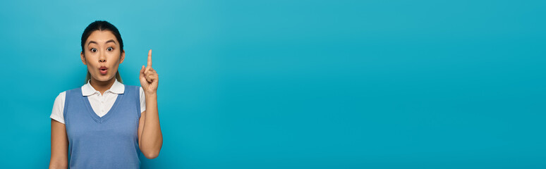 A young Asian woman in smart casual attire stands against a blue background, pointing upward with a...