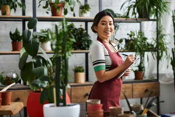 A young woman in an apron stands in her plant shop, taking inventory and surrounded by lush greenery.