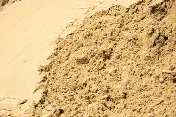 A close-up image of a pile of light brown sand, likely taken outdoors on a sunny day. The sand appears to be dry and loose, with a coarse texture