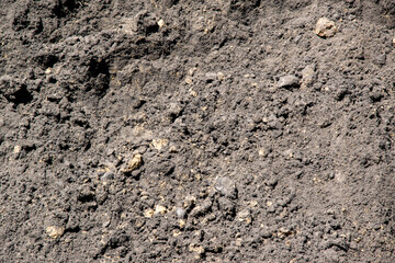 A close-up photograph of dark brown soil with small stones mixed in. The soil appears to be moist and loose
