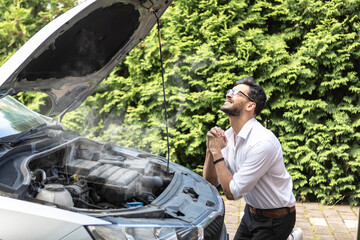 Shocked businessman stands in front of a broken car, opened the hood and tries to fix it	