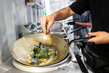 A person cooking mussels in a large wok, adding herbs for seasoning