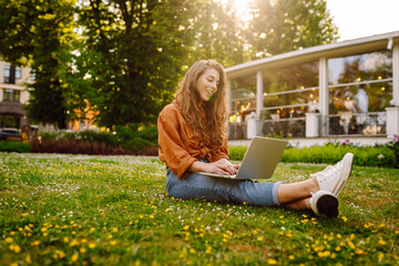 Young woman sits in park communicating by video call. Girl speaking looking at laptop computer,...