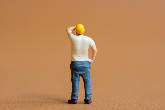Miniature construction worker. He stands with one hand on his hip while wiping sweat from his forehead