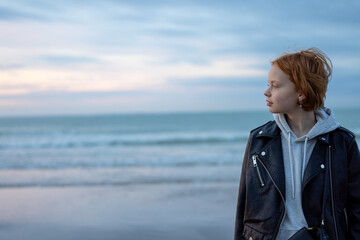 A young woman with red hair in a leather jacket and hoodie stares at the ocean, emanating calm and...