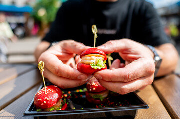 Close-up of hands holding a mini burger with colorful toppings, served on a black plate.