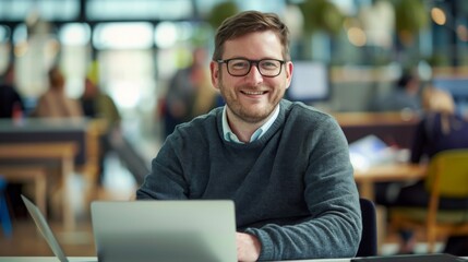 Fototapeta premium Smiling man in late thirties with glasses and short beard working on laptop at office desk, surrounded by busy modern open space environment, captured with wide angle lens