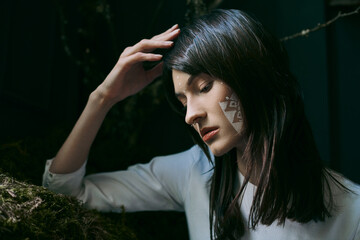 A woman with white face paint stands near a swamp.