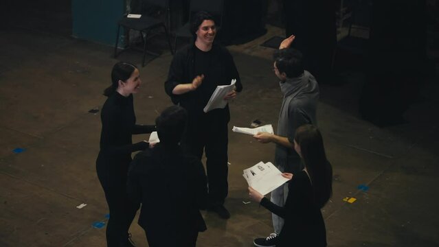 A happy and cheerful group of actors in black suits together with their stage director in a gray T-shirt and scarf are holding sheets of paper with a script in their hands and are ready to go on stage