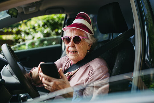 Happy senior woman driving car alone, holding smartphone and scrolling. Safe driving for elderly adults, older driver safety.
