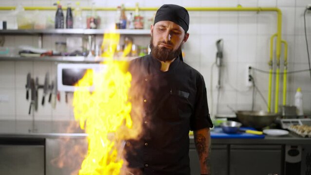 Confident male chef with a beard uses the flambeing technique while frying vegetables