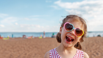 Banner laughing cheerful girl 5 years old in heart-shaped sunglasses with cherry in your mouth on beach in summer day