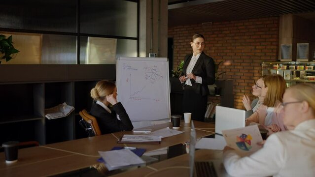 A young woman with brown hair wearing round glasses and a professional attire is pitching her idea at a stand during an office meeting