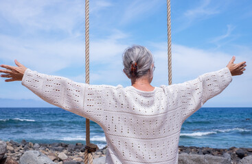Back view of senior woman with outstretched arms sitting on swing on sea beach enjoying relaxed and fun moments, retirement lifestyle for an elderly lady in summer vacation