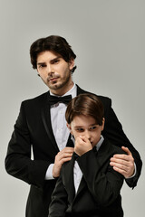 A father in a tuxedo calmly reassures his son, who is also in a tuxedo, with a hand on his shoulder.