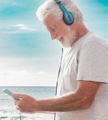 Smiling bearded senior man holding mobile phone sitting in outdoors at the beach listening music by earphones, elderly male enjoying  vacation, free time and retirement