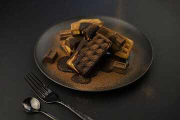 World Chocolate Day. pieces of chocolate and waffles sprinkled with cocoa powder on a black plate and dark background.