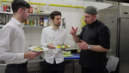 Two male waiters in white shirts communicate with a chef in a black uniform about the quality of food while standing in the kitchen in a restaurant. A professional chef in a black uniform tells the