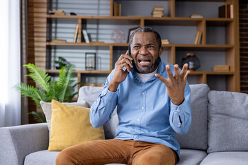 Frustrated man holding a phone while sitting on a sofa. He looks upset and is gesturing with his hand in a modern living room.