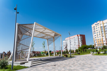 A serene urban landscape features a peaceful white building nestled in a park with lush greenery and city buildings in the background. The scene creates a tranquil ambiance