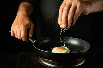 Add salt by hand to a hot frying pan with eggs. Chef preparing egg healthy breakfast in restaurant kitchen