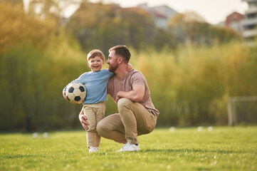 Sitting, holding soccer ball. Happy father with son are having fun on the field at summertime