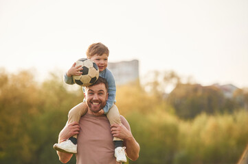 On shoulders, with soccer ball. Happy father with son are having fun on the field at summertime