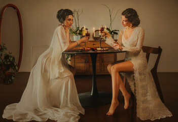 Two women Sexy girl beauty face lips sitting at the dinner table luxury light room. vintage nightie silk white long dress fairy old style noble lady girls holds glass wine drink clink glasses toast