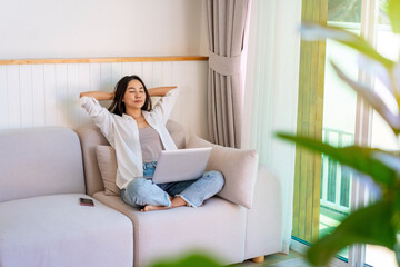 Young Asian woman relaxing and using laptop on a sofa at home in the morning
