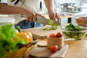 A cropped image of a woman slicing a cucumber with a knife on a cutting board at a kitchen table, cooking in the kitchen. healthy food, home cooking, wellness eating, domestic life
