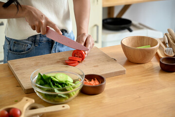 A beautiful, happy young Asian woman cutting a tomato on a cutting board, preparing ingredients for her healthy salad, enjoying cooking in the kitchen. home cooking, domestic life, healthy eating