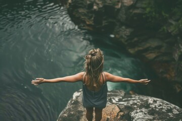 Fototapeta premium A woman is standing on a rock near a body of water, with her arms outstretched