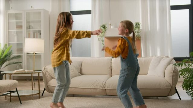 Two carefree Caucasian children little girls sisters laugh jumping floor celebrating moving bouncing kids friends daughters dancing music having fun together family home apartment friendship housing