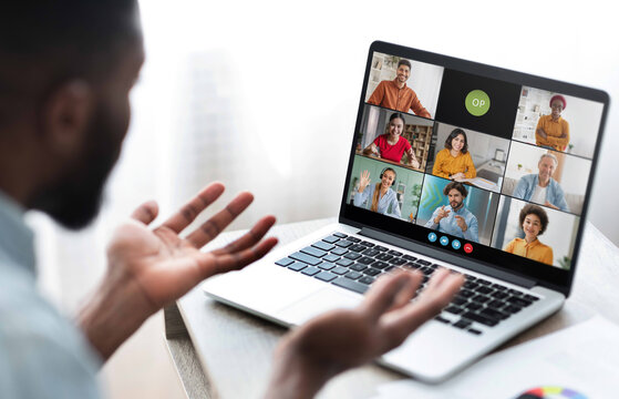 Black man sits in front of a laptop, participating in a video conference meeting with several colleagues. He has his hands raised as if he is making a point during the call.