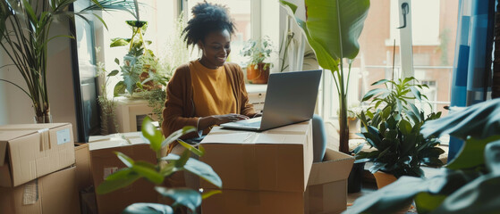 Woman working on laptop amidst moving boxes and an array of indoor plants, representing a blend of work and home relocation.