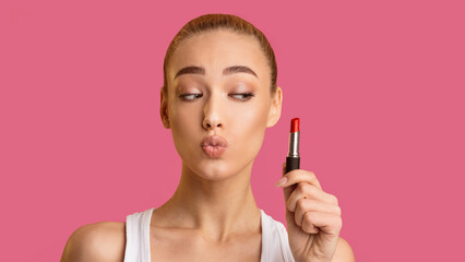Fototapeta premium A young woman with blonde hair is holding a red lipstick in her right hand and is about to apply it to her lips. She is pouting her lips and looking at the lipstick. The background is a solid pink