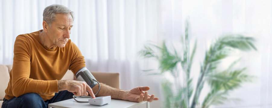An elderly man wearing a yellow sweater sits at a table in his living room and uses a digital blood pressure monitor to check his blood pressure, copy space