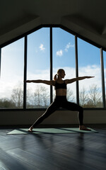 Woman is practicing ashtanga yoga vinyasa flow, warrior two asana pose exercise indoors, against the window. Wellness, lifestyle, copy space.