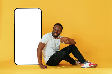 Sitting near to the large smartphone with copy space. Black man is in the studio against yellow background