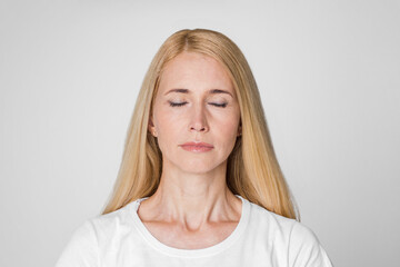 Fototapeta premium A close-up photo of a blonde woman with closed eyes, standing against a simple white background. She appears to be meditating or in a peaceful state.