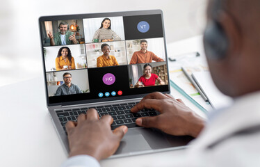 A man sits at a desk, his hands typing on a laptop keyboard, while participating in a video conference. The screen shows six participants in individual video frames.