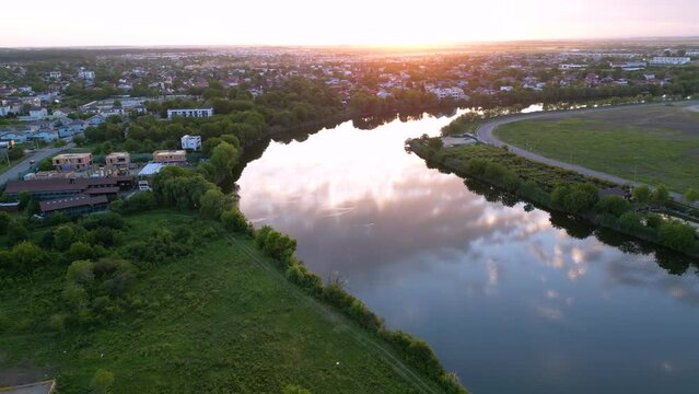 Aerial sunset view of a calm river winding through a low-rise urban and suburban landscape, with residential neighborhoods, green riverbanks, roads and open development land reflecting warm evening li