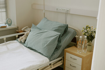 Hospital ward with single bed with light mint linen and wooden night stand with vase full of daisies and two bottles of water on top