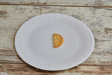 One half of a galette cookie cookie on a white disposable plate on a wooden background close-up, lack of food, rising food prices, meager diet, food shortages