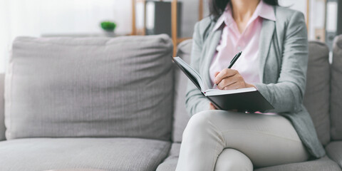 A woman sits on a gray couch in a living room. She is writing in a black notebook with a pen in her right hand. The scene is calm and serene, with the woman focused on her writing, copy space