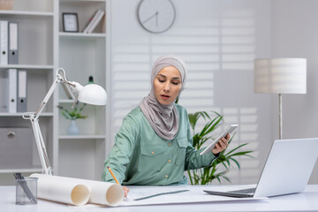 Female architect in hijab working on project using tablet and laptop in a contemporary office setting.