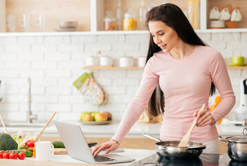 A woman is multitasking in the kitchen, cooking food while using a laptop for work or browsing the...